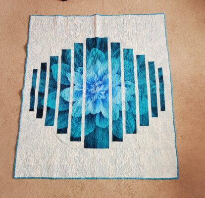 Intricately quilted work featuring a large blue flower divided into vertical strips.