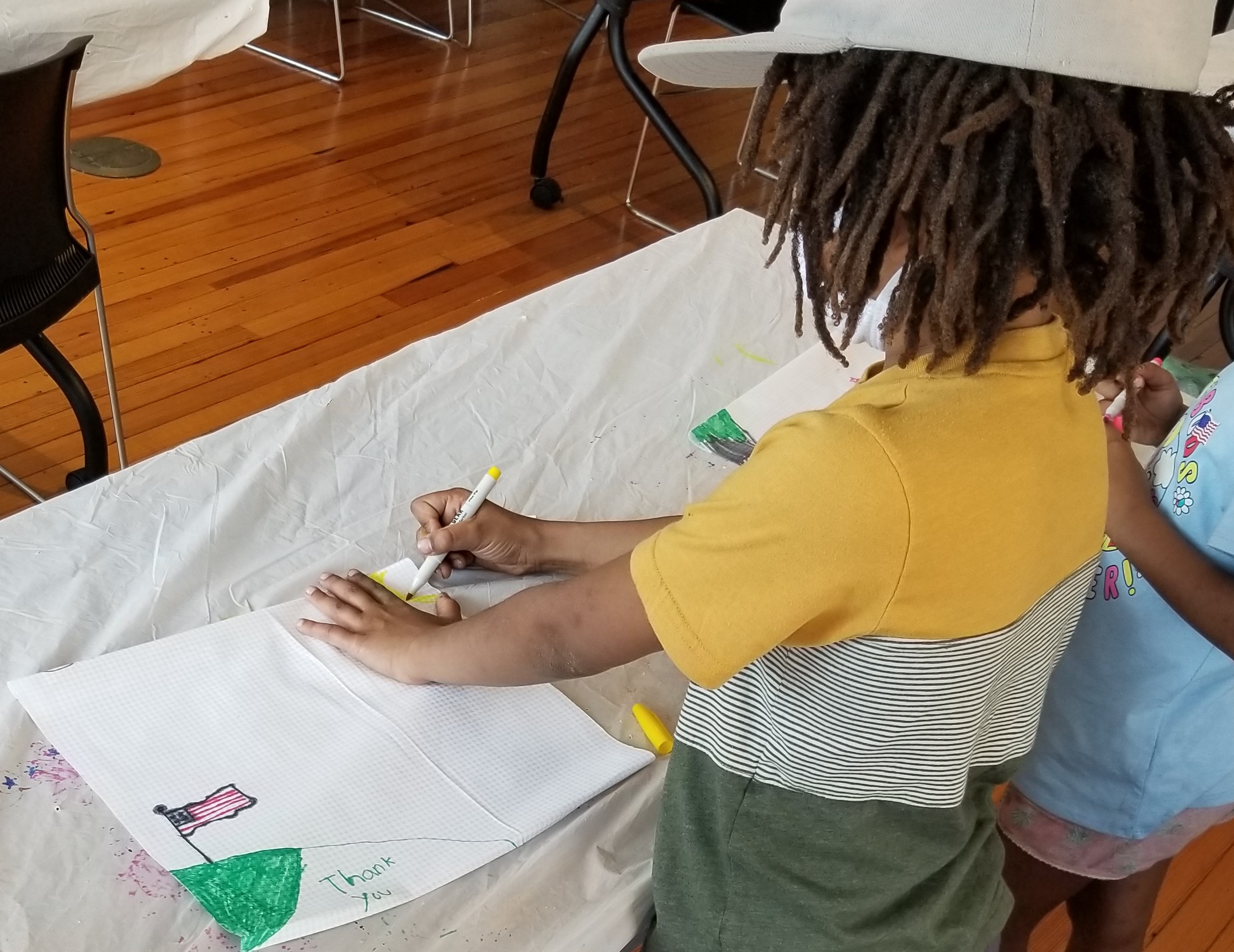 A BIPOC-presenting child draws a picture showing an American flag and the words "Thank you."