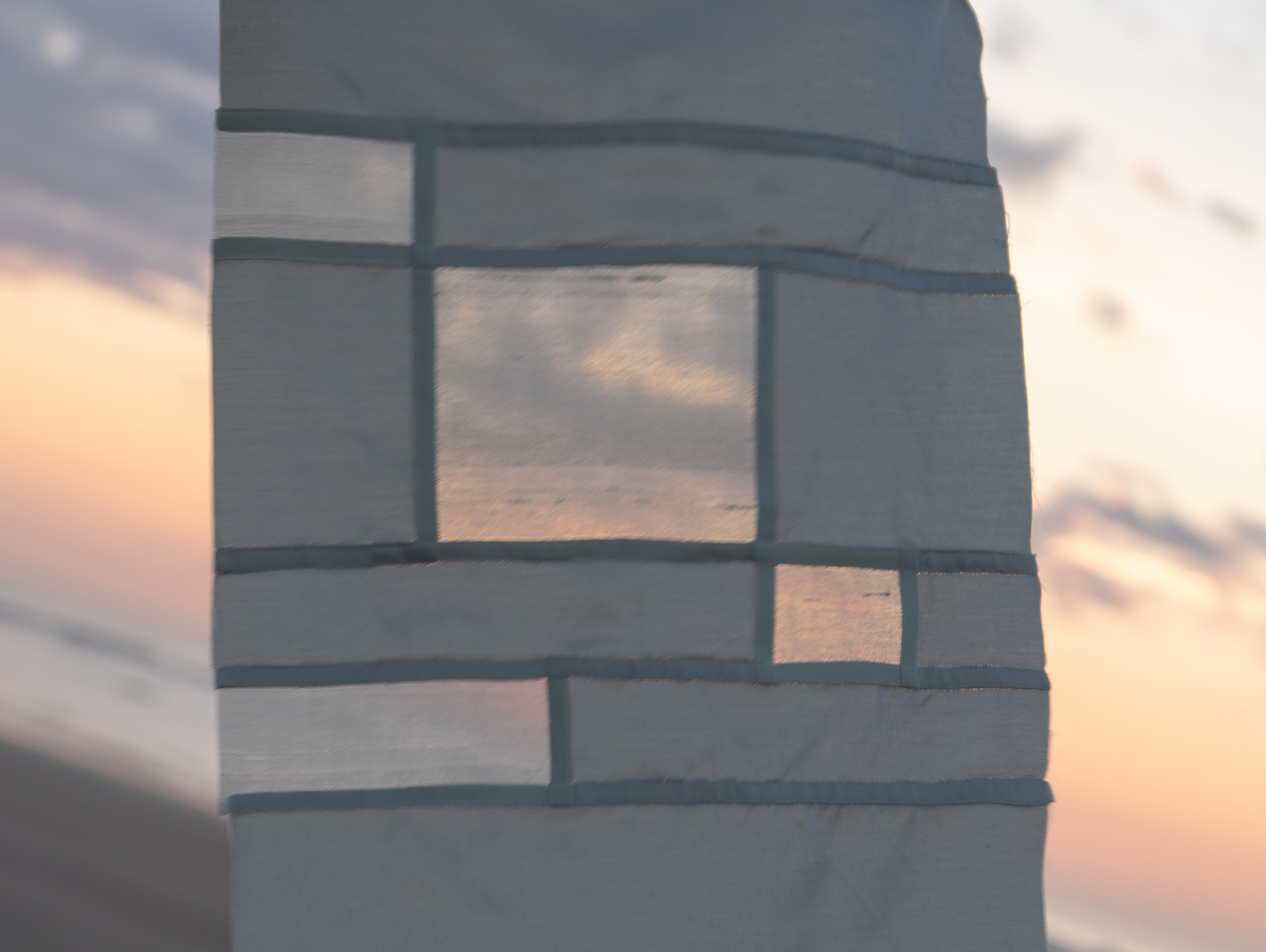 A small, gray patchwork bojagi panel of fabric, held up against a sunsetting sky.