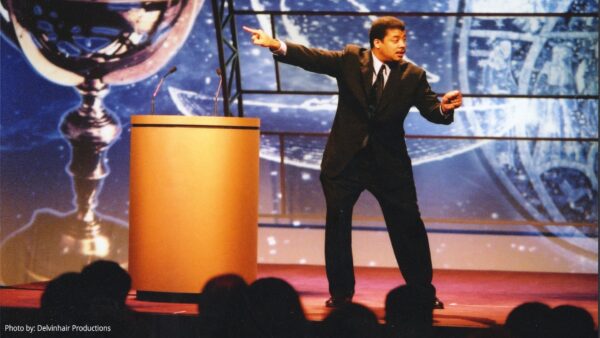 A black man in a dark suit gestures as he speaks to an audience from a lit stage with a podium.
