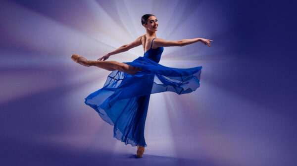 A smiling ballet dancer in arabesque en pointe, wearing a blue dance dress with a flowing skirt.