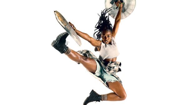 A Black dancer mid-leap, wearing combat boots and holding cymbals.