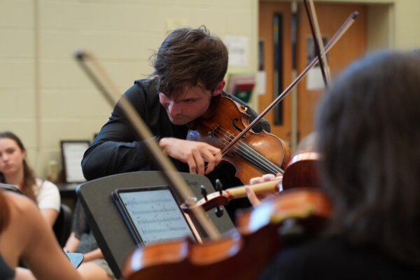 A male violinist plays, standing, among a youth orchestra.