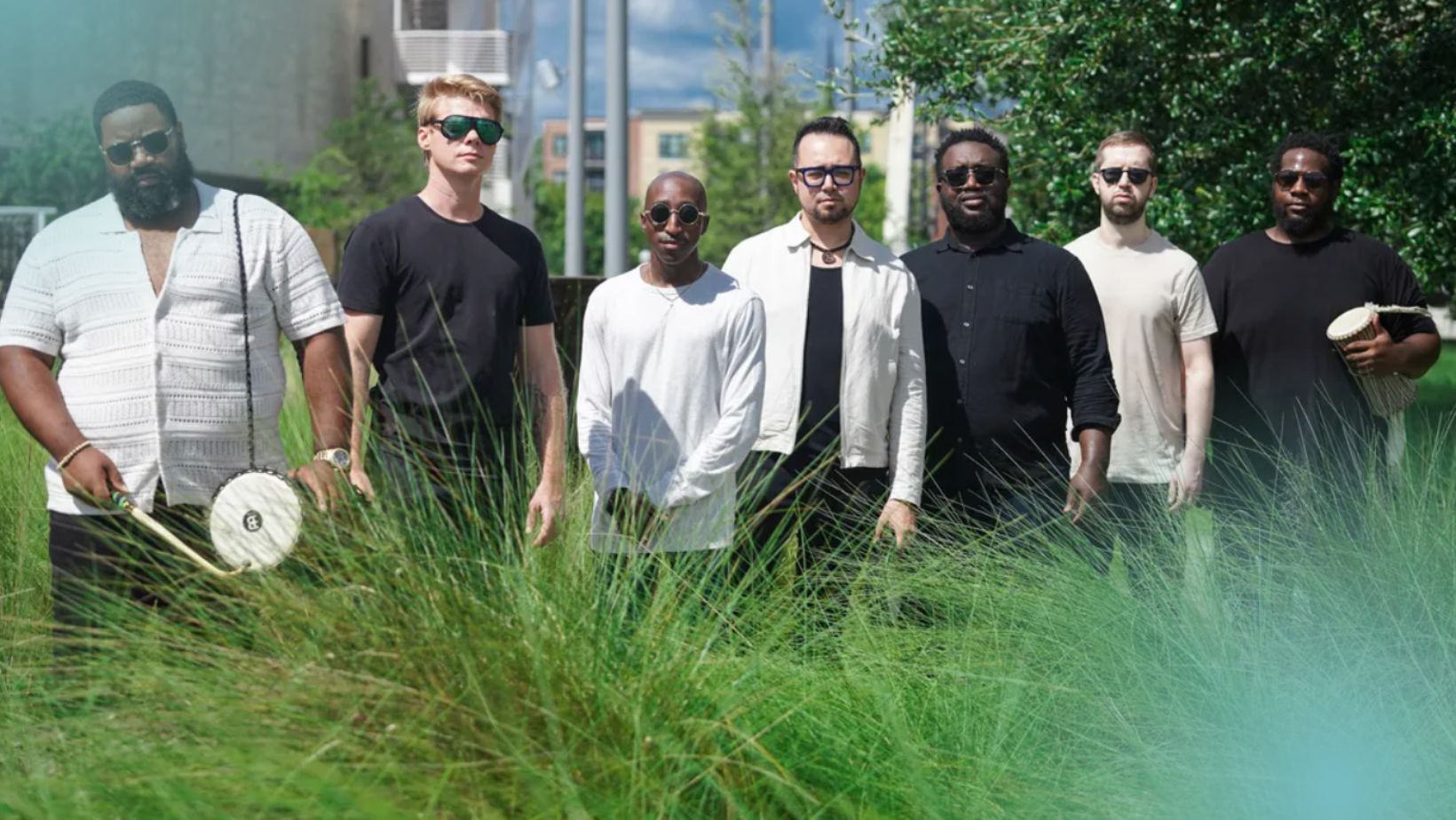 7 men stand with small drums amid tall grasses outdoors. 4 present as Black.