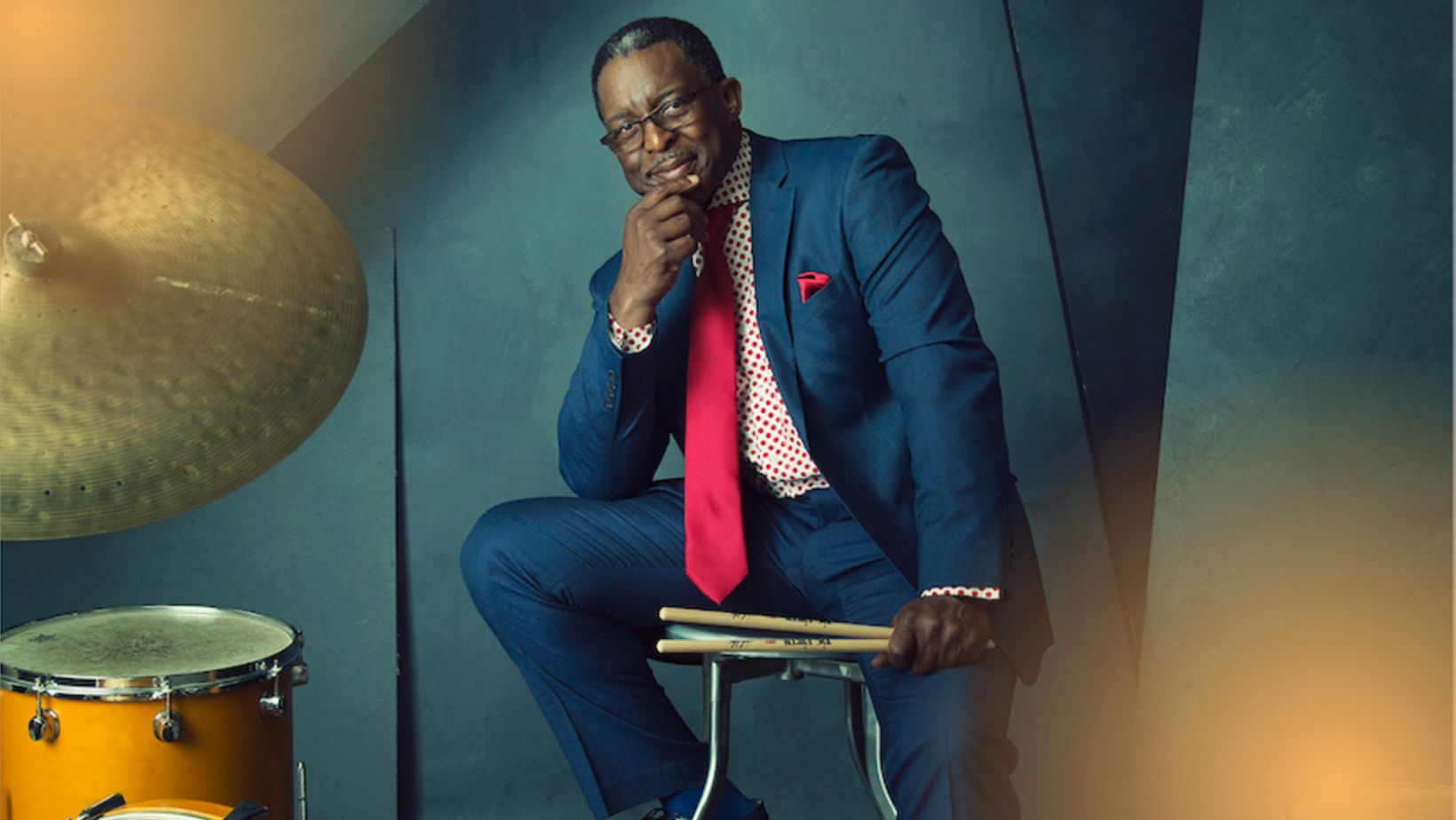 A Black man in a blue suit sits with a drum set. One hand cups his chin and he smiles slightly.