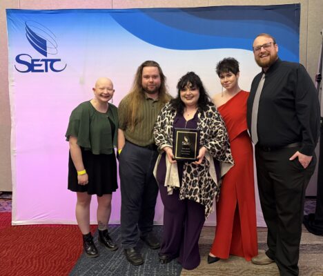 Five people stand in front of a blue and white SETC banner. The central person holds the award plaque.