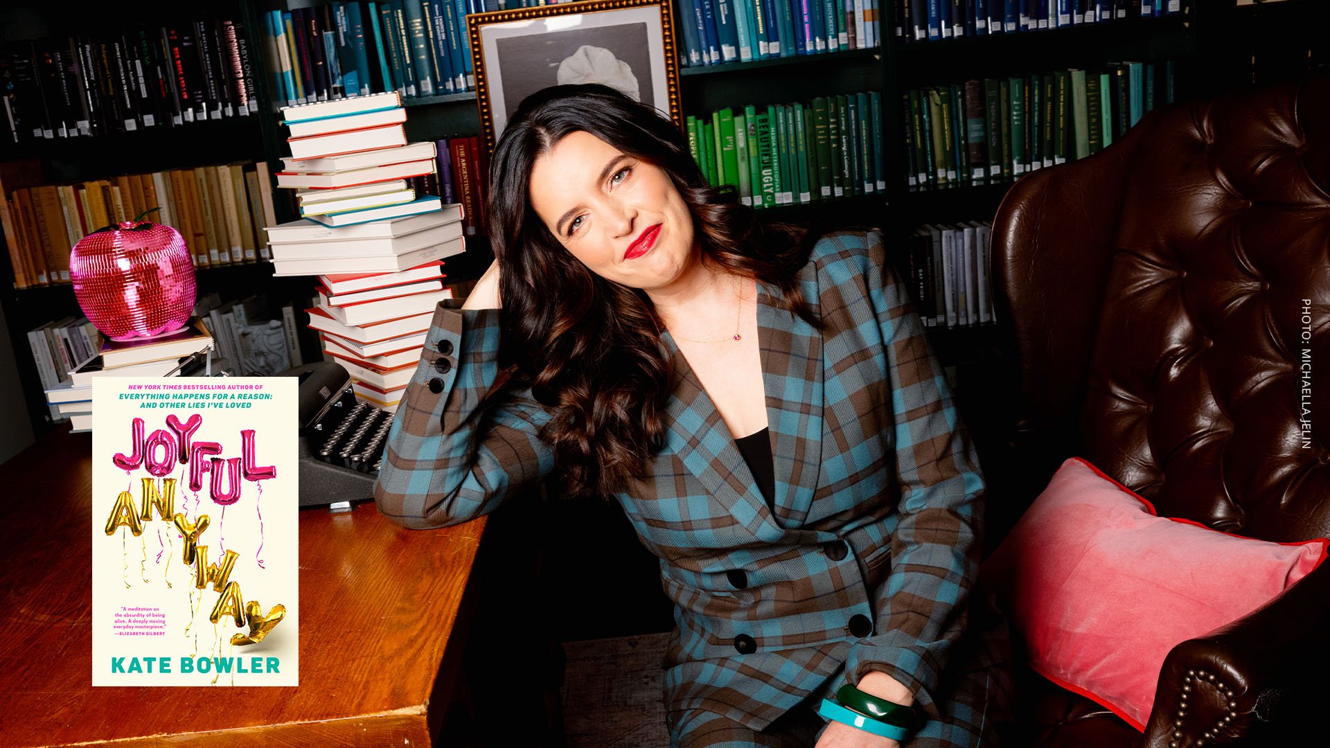 Woman with long dark hair and bright lipstick sitting amid books, including her own, Joyful Anyway.