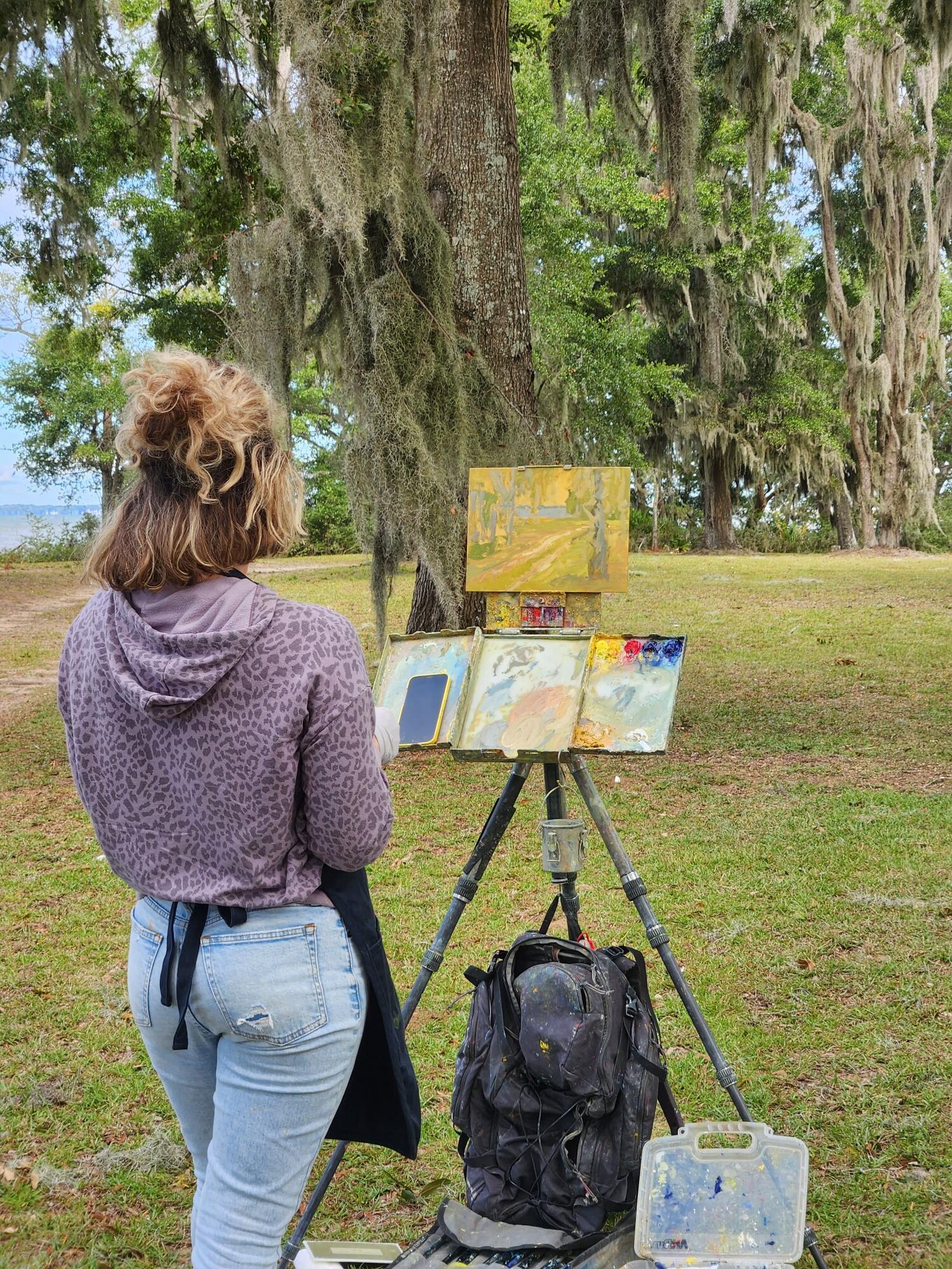 A female painter at an easel in a grassy area with mature, mossy trees.
