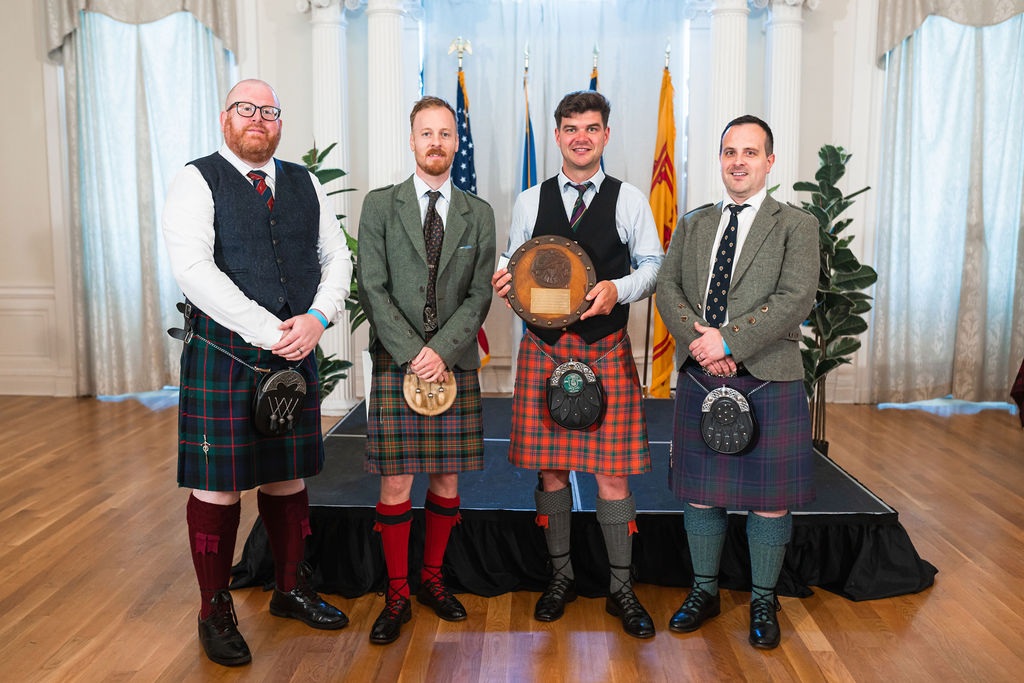4 men in Scottish garb, including kilts, pose for an indoor photo. One holds a traditional drum.