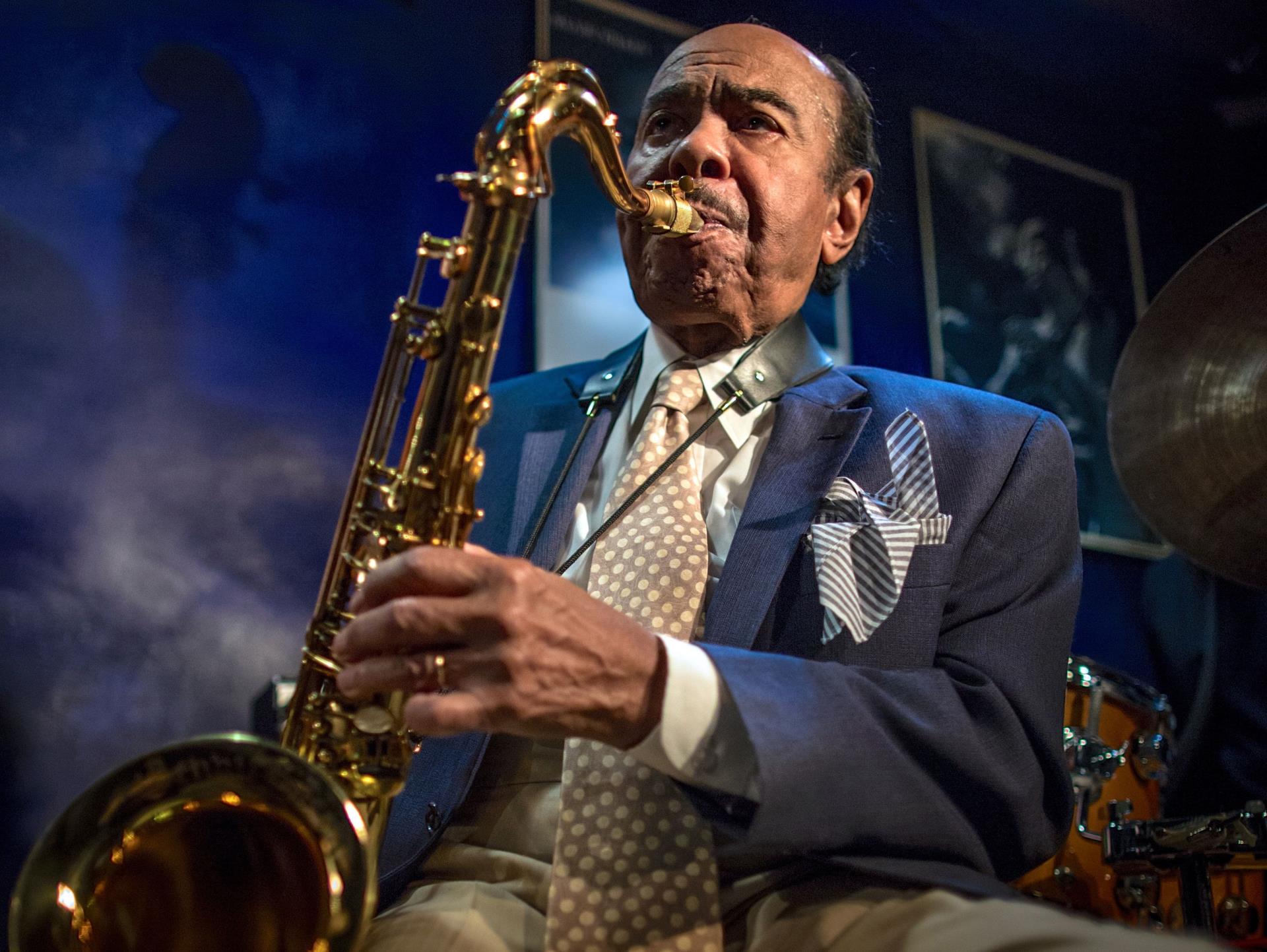 A seated Black man plays saxophone. He has a thin mustache and a receding hairline.