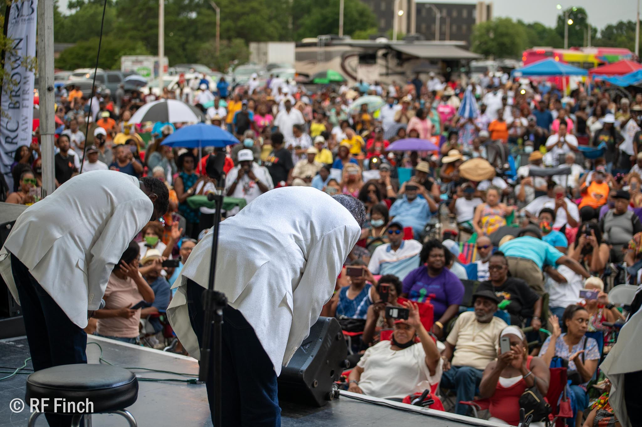 2 men in white suit jackets and black pants bow deeply onstage for a large outdoor audience.