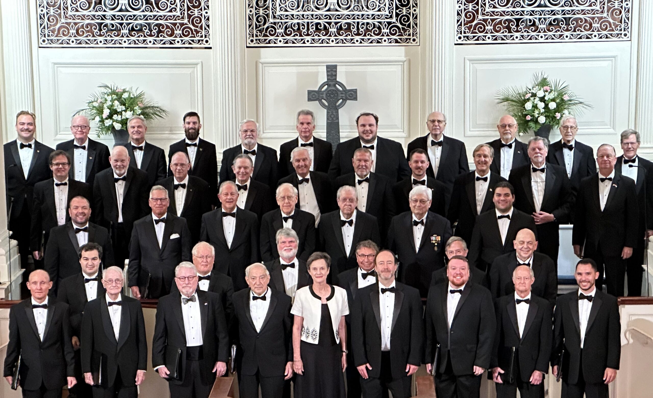 A choir of about 40 men in tuxedos stand ready to sing in a church.