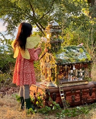 A young girl wearing a dress and butterfly wings stands at a miniature house in a wooded area.