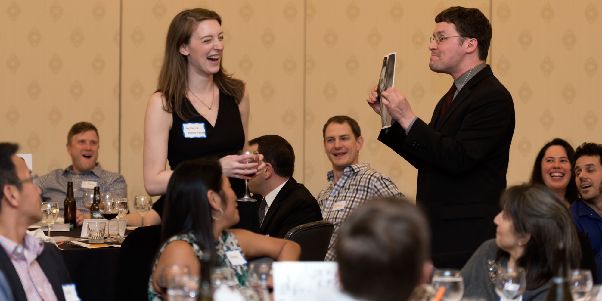 A standing woman laughs as a standing man shows her a photo amid a seated audience of spectators.