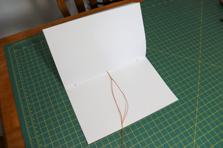 Folded paper on a gridded work surface, with a needle and thread for bookbinding.