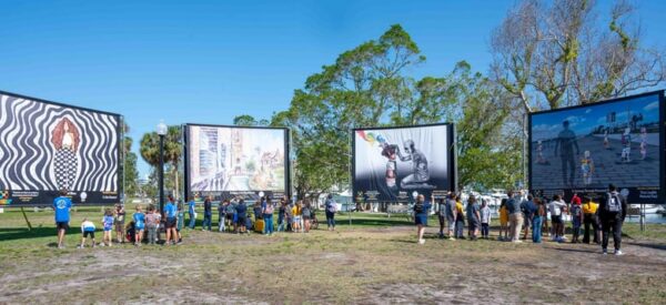 Groups of people of all ages look at artwork displayed on 4 billboard-sized panels in an open field.
