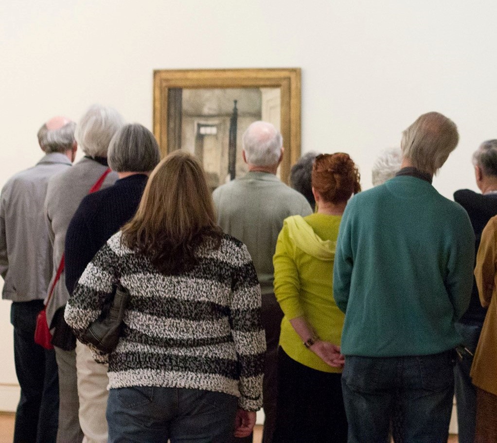 A group of people seen from behind, viewing a painting in a museum.