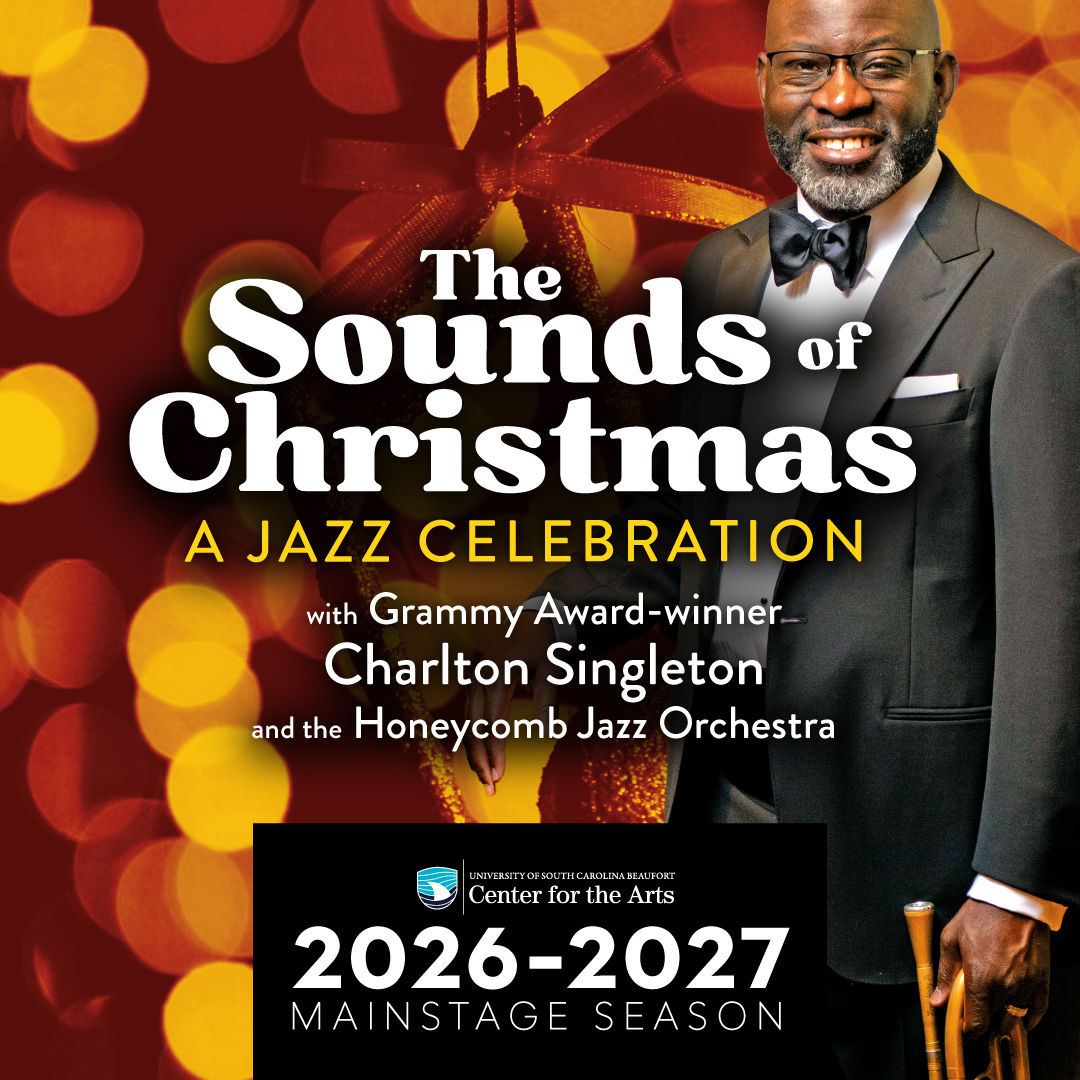 A Black man in a tuxedo holds a brass instrument at his side, against a background of lights.