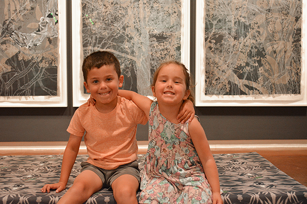 Two children sit in front of large artworks. They are smiling.