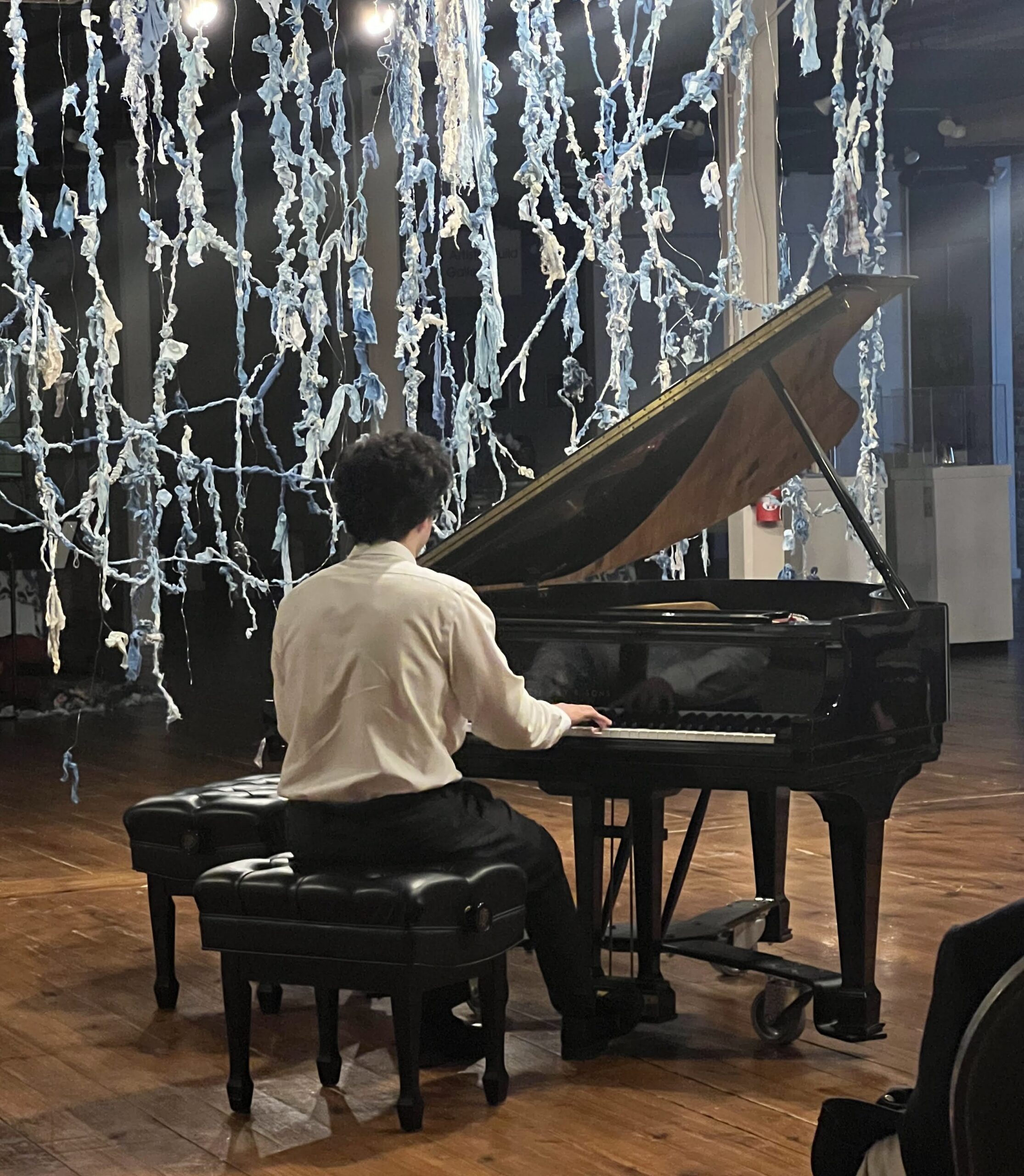 A person plays a baby grand piano in a room with a fabric art installation.
