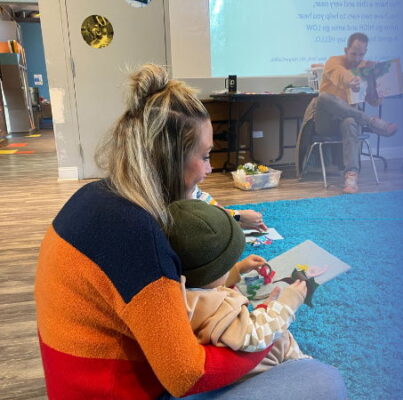 A blonde woman holds a small child in her lap, reading to them.