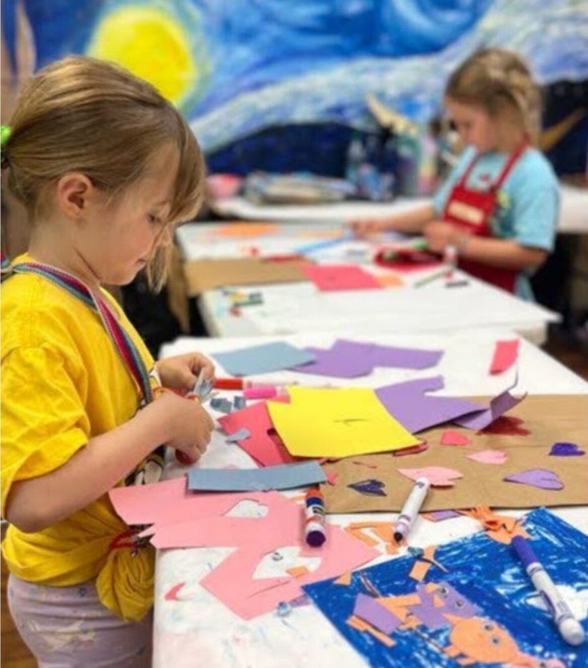 Children wearing aprons make art with construction paper and markers on a work table.