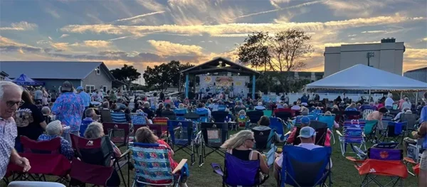 Green space filled with people in chairs attending an outdoor performance at sunset.