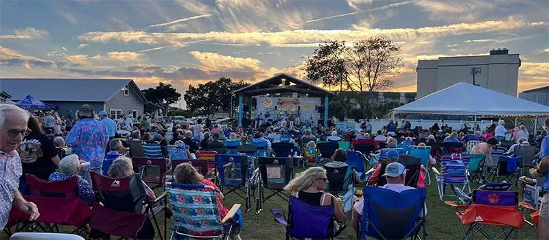 Green space filled with people in chairs attending an outdoor performance at sunset.