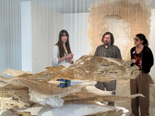 A male and two females look at an installation artwork in an indoor gallery.