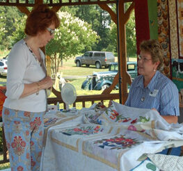 An artisan with a handmade quilt draped on a table engages an Ag and Art Tour visitor about the craft. The are on a covered porch on a sunny day.